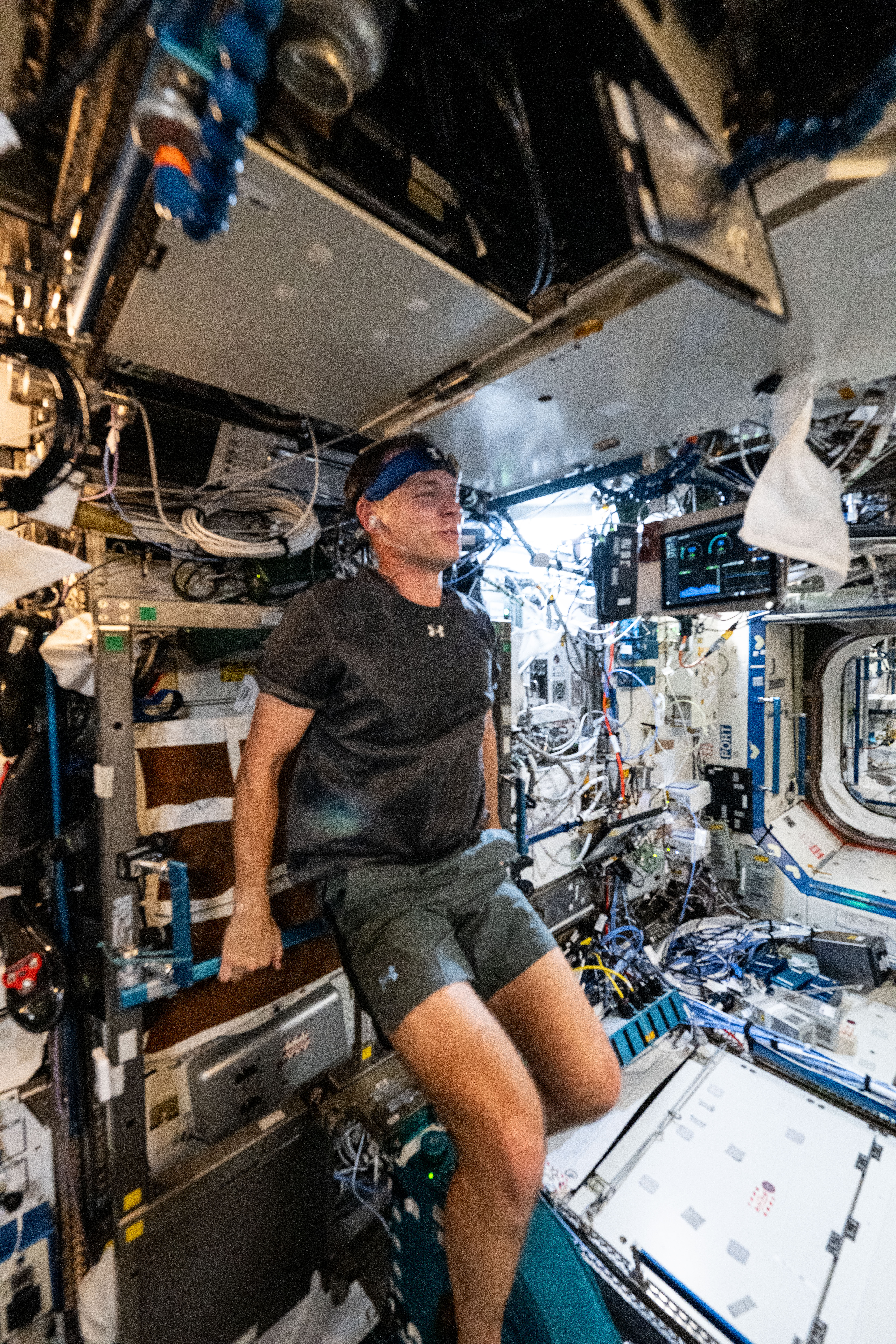 NASA Astronaut Nick Hague is wearing a gray t-shirt and gray shorts, holding a white towel in his left hand. He is facing away from the camera and smiling.