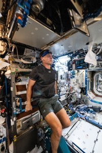 NASA Astronaut Nick Hague is wearing a gray t-shirt and gray shorts, holding a white towel in his left hand. He is facing away from the camera and smiling.