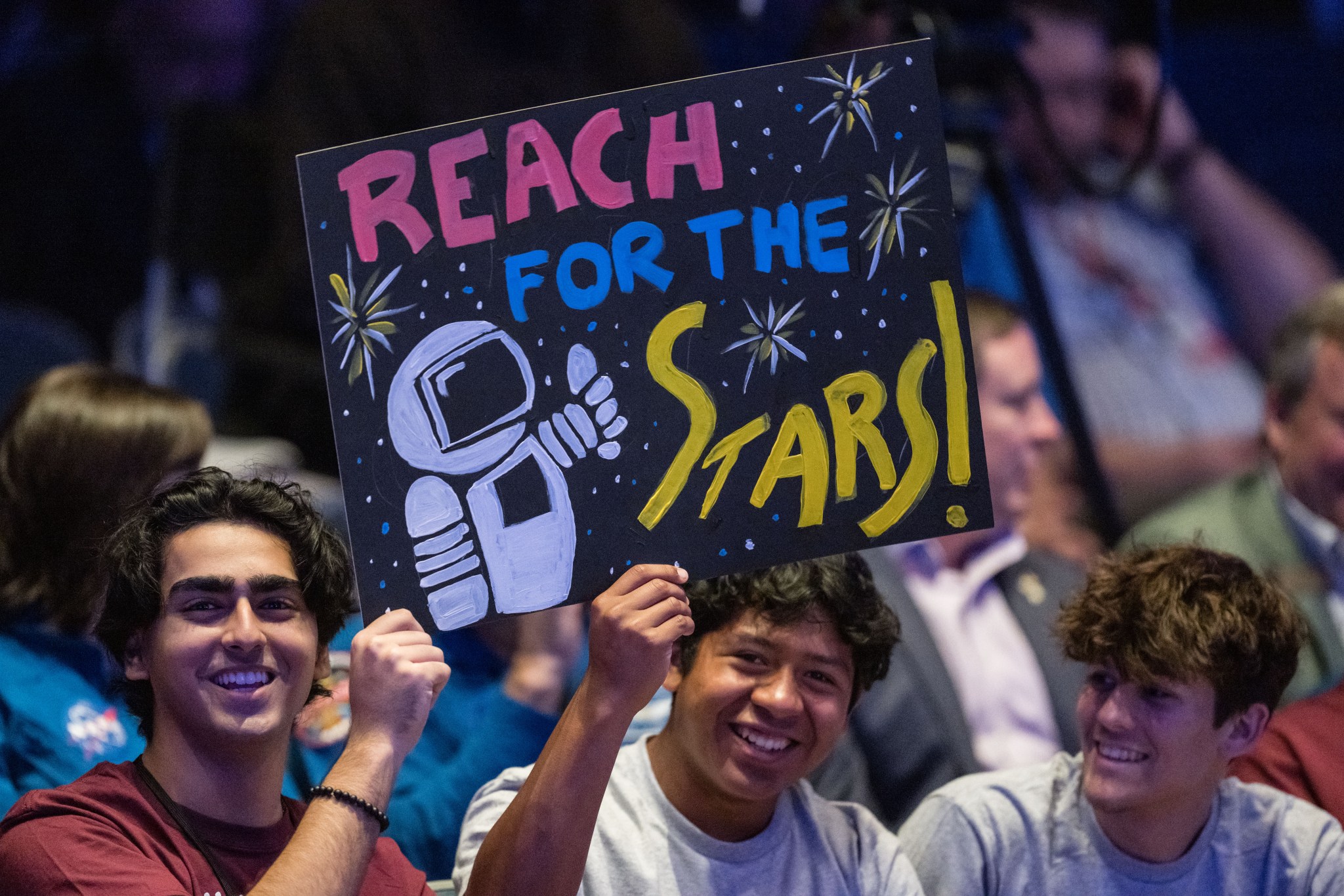 A group of three male interns congratulate the 23rd class of astronauts at NASA's Johnson Space Center in Houston on March 5, 2024. The trio smile while holding a colorful sign displaying an astronaut giving a thumbs up and text reading “Reach for the stars!” The group wears casual attire and sit before an audience of other members of the NASA workforce and community. Launch Your Career with NASA. 