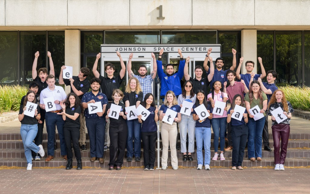 A group of young people stand in front of NASA’s Johnson Space Center holding signs that spell out ‘#BeAnAstronaut.’ The participants are raising their arms and smiling.