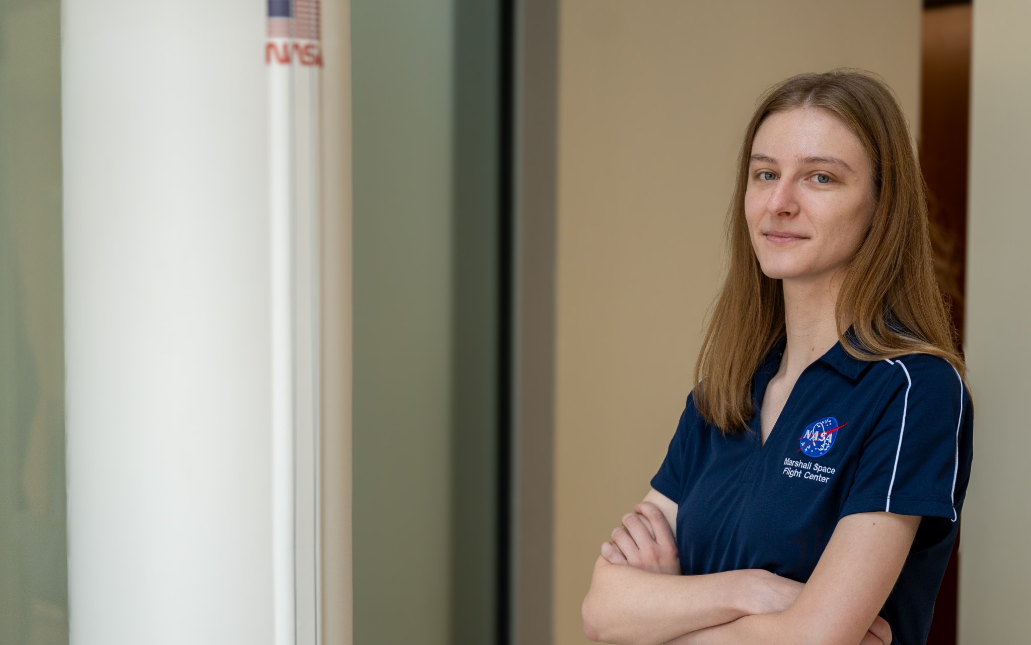 NASA Human Landing System Program Raptor engine insight lead Sarah Ryan poses in front of a model of SpaceX’s Starship human landing system. Sarah is in a navy shirt and khaki pants. She poses with her arms crossed in front of her chest. The black and white lander is just over her right shoulder.