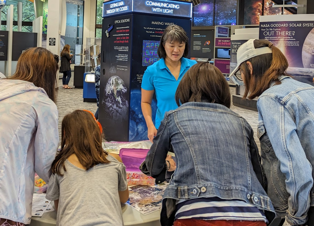  A woman wearing a blue polo demonstrates an activity on a table in front of four participants at the Goddard Visitor Center