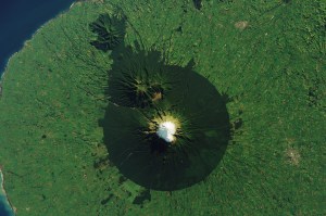 An overhead view of Mount Taranaki from a satellite. A white peak (a snow-capped mountain) is circled by dark green forest. The rest of the land around it is a bright green.