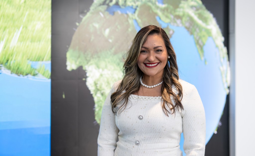 Yaíta smiles broadly at the camera wearing a white dress with a pearl necklace. Her hair is long with curls and she's wearing red lipstick. She stands in front of a larger model of a globe.