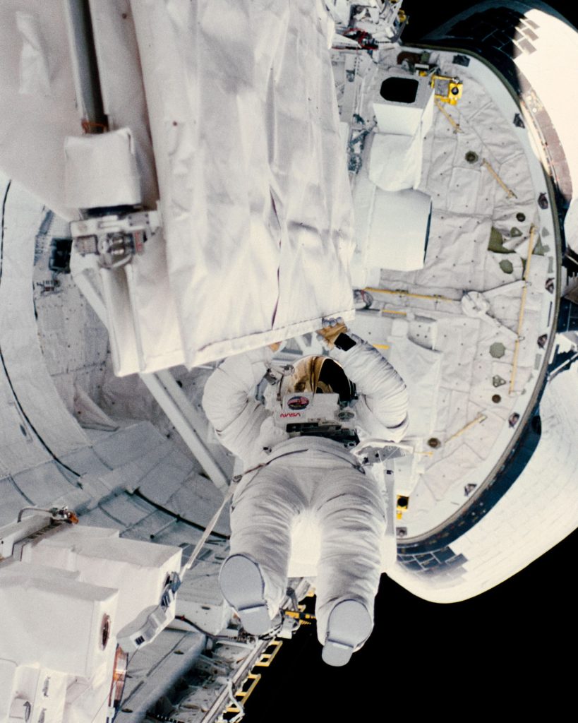 Astronaut Kathryn D. Sullivan checking the latch of an antenna in the space shuttle Challenger’s open cargo during her historic extravehicular activity
