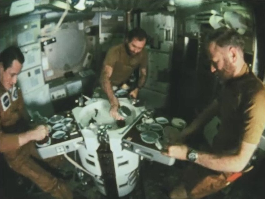 Three astronauts in brown shirts are positioned around a table are eating a Thanksgiving meal together. The astronauts are surrounding by the interior walls of NASA's Skylab. The walls are white with multiple drawers.
