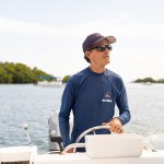 A Puerto Rican man wearing sunglasses, a navy baseball cap and longsleeve shirt is visible from the waist up, standing behind the silver wheel of a small white boat. In the background are a few islands of green foliage.