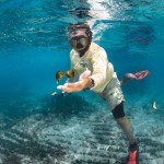 A Puerto Rican man wearing a snorkel mask, fins, and a cream long-sleeve shirt float in bright blue water with one hand extended towards the camera. Just above his hand float two brown fuzzy balls, roughly the size of golf balls.