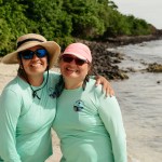Two Puerto Rican women stand on a beach, both wearing teal longsleeve shirts and sunglasses. The one on the left has a tan, wide-brimmed hat, and the one on the right has a pink baseball cap.