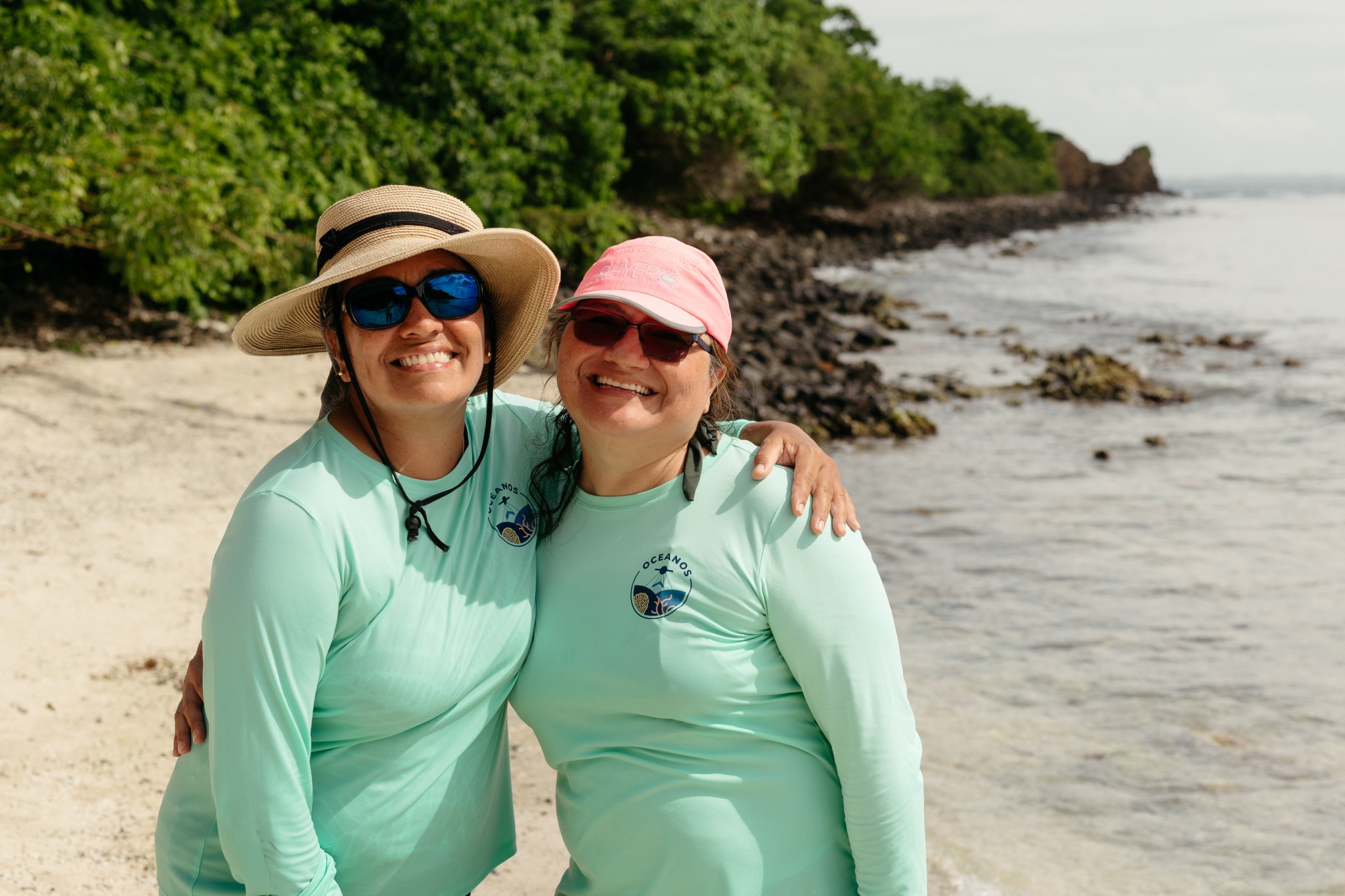 Two Puerto Rican women stand on a beach, both wearing teal longsleeve shirts and sunglasses. The one on the left has a tan, wide-brimmed hat, and the one on the right has a pink baseball cap.