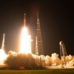 An orange rocket lifts off from a launchpad. The sky is black, but the fire coming from the engines lights up the surrounding area, highlighting clouds of white vapor spreading across the ground.