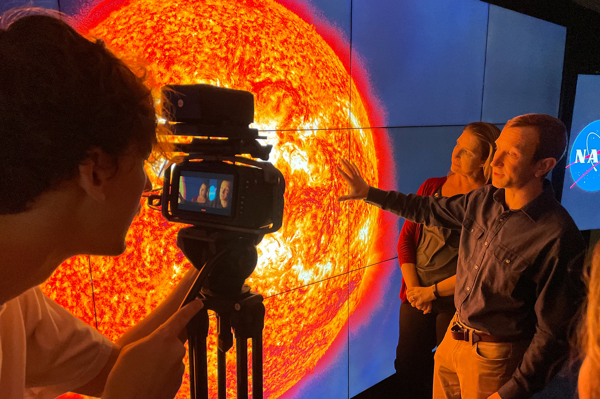 A man stands in front of a multi-paneled screen, which shows a large image of the Sun. A woman in the background looks on while a person at the left of the frame records the man with a video camera.