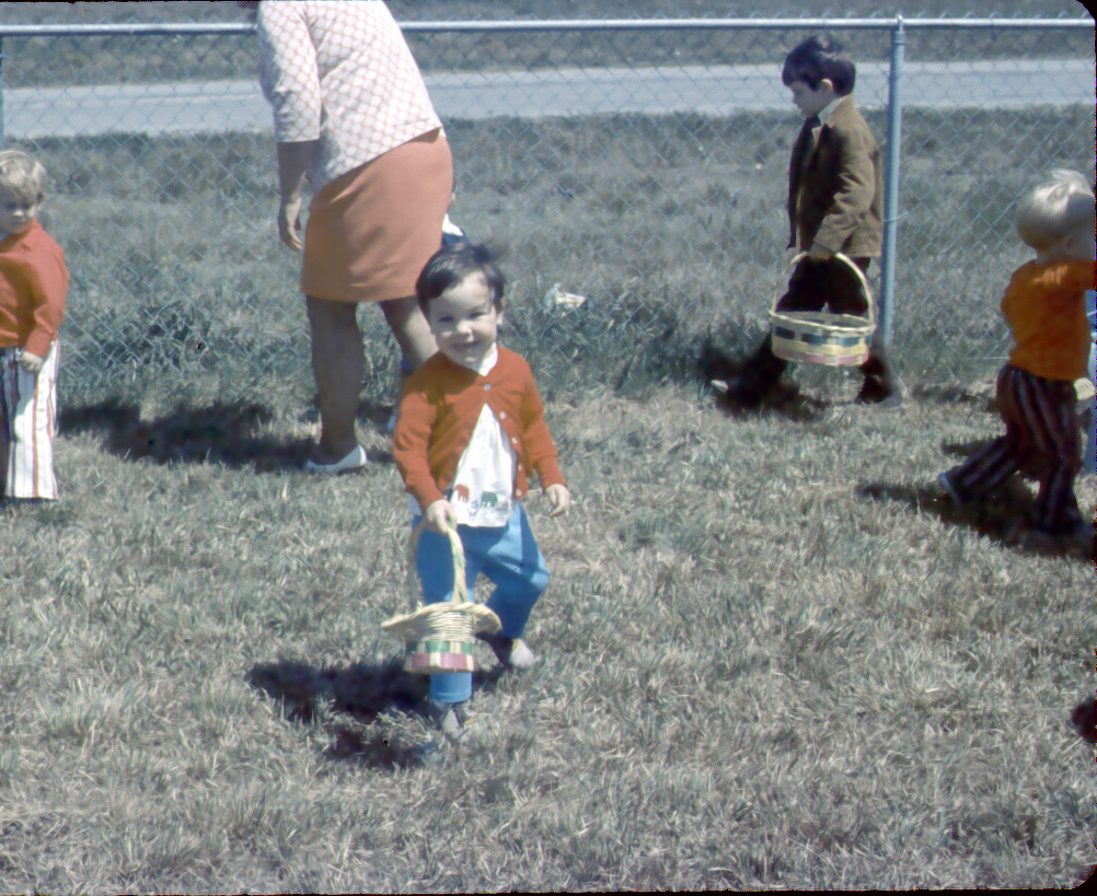 Children participate in an outdoor Easter egg hunt on a grassy field near a chain-link fence. A young child in the foreground holds a small basket.