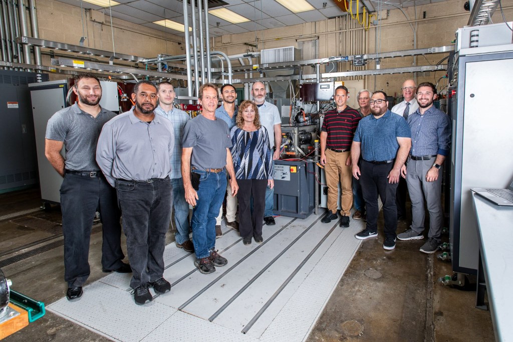 A group of 12 people pose in front of machinery and equipment inside a testing room supporting research for vertical lift vehicles and sustainable transportation.