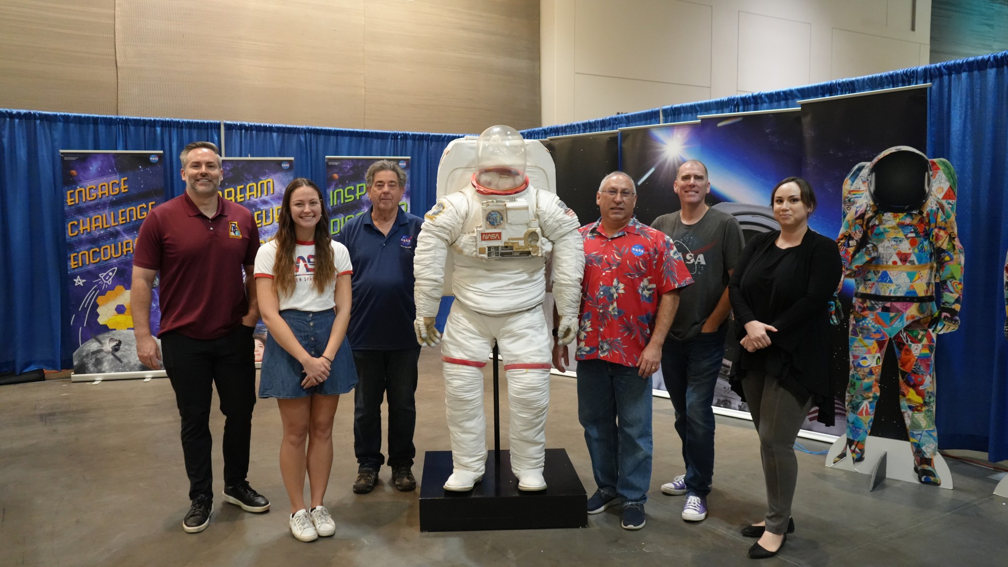 A group photo at a NASA booth features six individuals standing alongside an astronaut suit display.