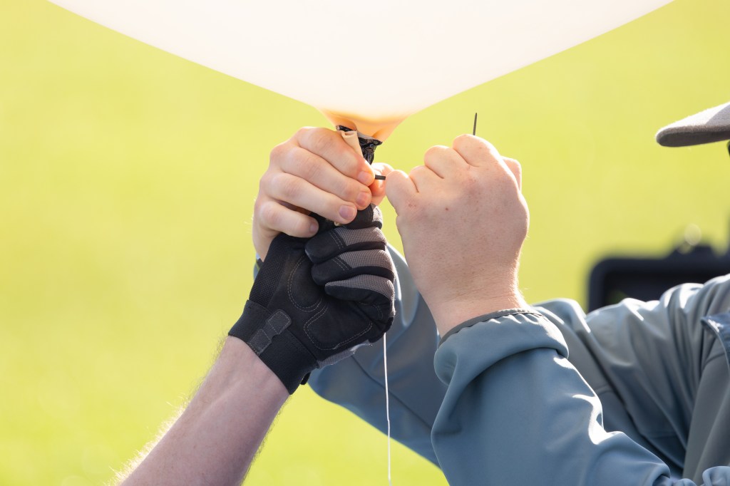 Logan Kearney and Cole Long, students at the University of Idaho, tie a zip tie to the base of a weather balloon as part of the preparations for launch, during FireSense’s uncrewed aerial system (UAS) technology demonstration in Missoula, Montana. These balloons use a sensor called a radiosonde to gather data on wind speed and direction.