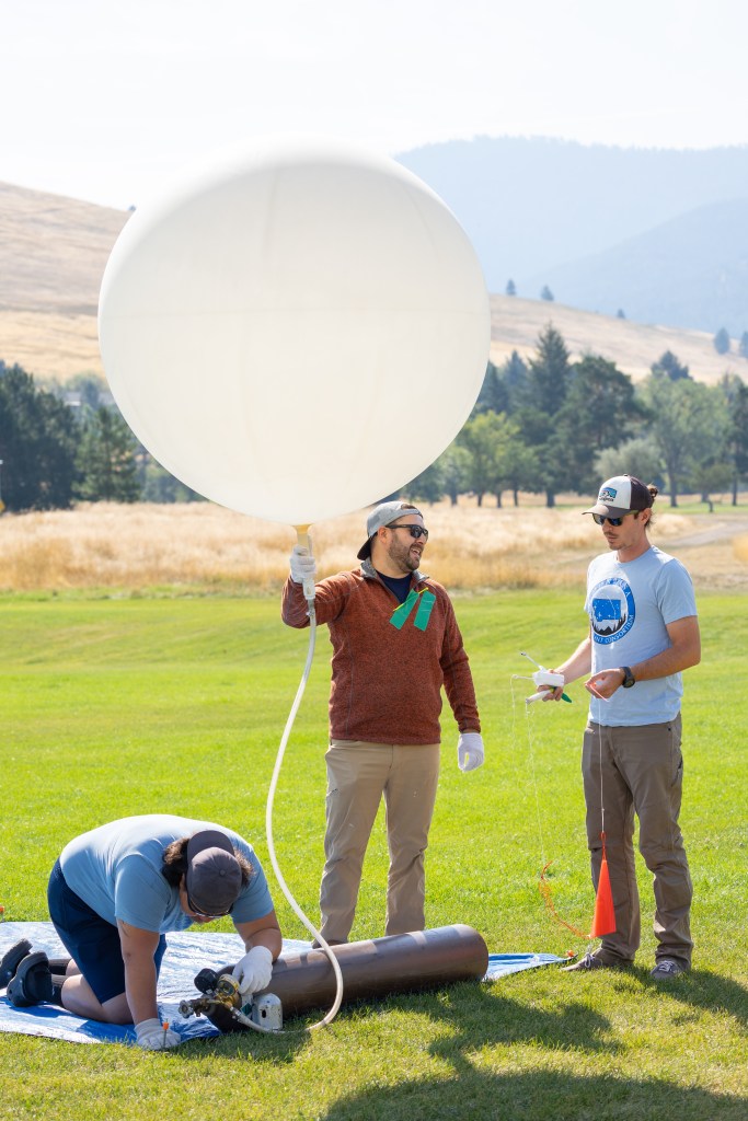 Steven Stratham (right, light blue shirt) attaches a radiosonde to the string of a weather balloon as teammates Travis Christopher (left) and Danny Johnson (center) prepare the balloon for launch. This team of three from Salish Kootenai College is one of many college teams across the nation trained to prepare and launch weather balloons under a Space Act Agreement, and were one of two teams that supported the FireSense UAS Technology Demonstration in Missoula, Montana.
