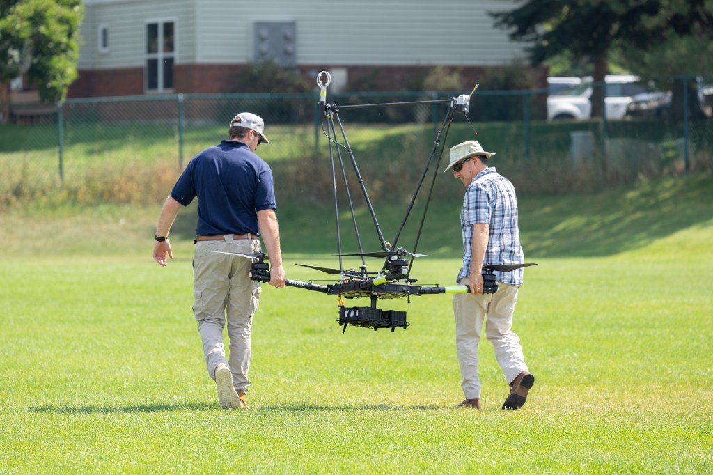 UAS Pilot in Command Brayden Chamberlain (left) and mechanical engineer Todd Ferrante (right) carry the NASA Alta X quadcopter into the field for a launch, during the FireSense uncrewed aerial system (UAS) technology demonstration in Missoula, Montana.