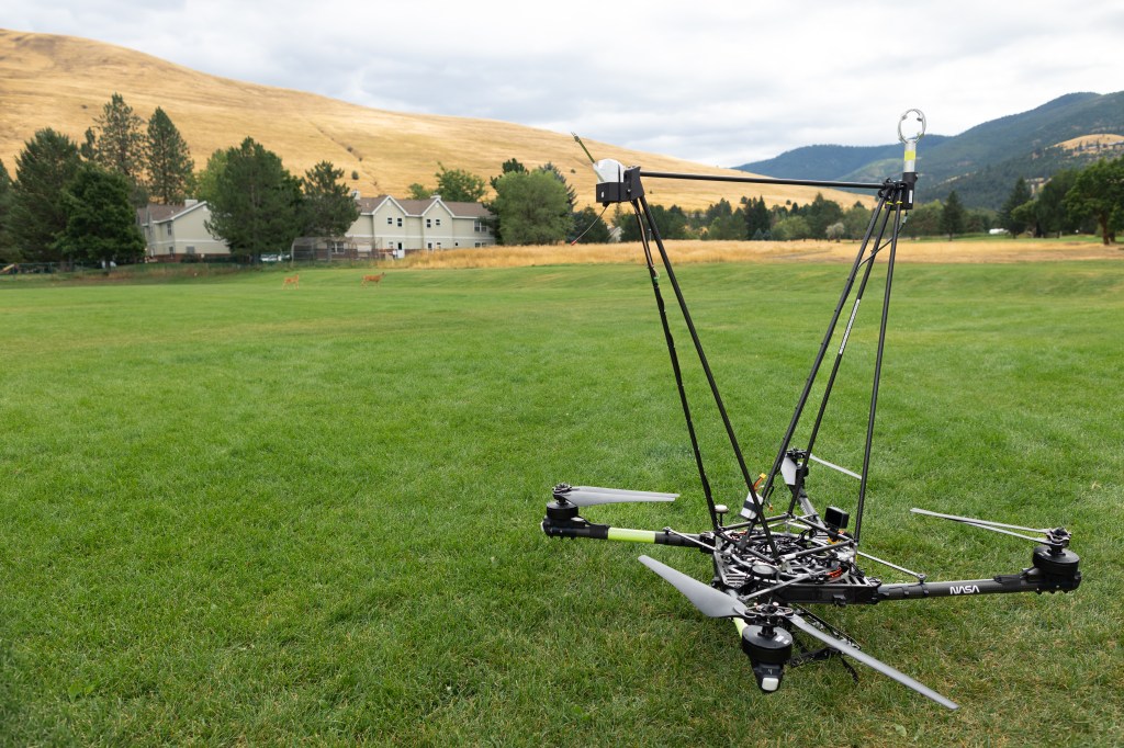 The NASA Alta X quadcopter sits in a field in Missoula, Montana, outfitted with a structure engineered at Langley Research Center to carry a radiosonde and an anemometer into the air. In the background, two deer make their way across the field. The drone and its payload were part of the August 2024 FireSense campaign, which looked at the applicability of using controllable, repeatable airborne measurements to more accurately predict fire and smoke behavior.
