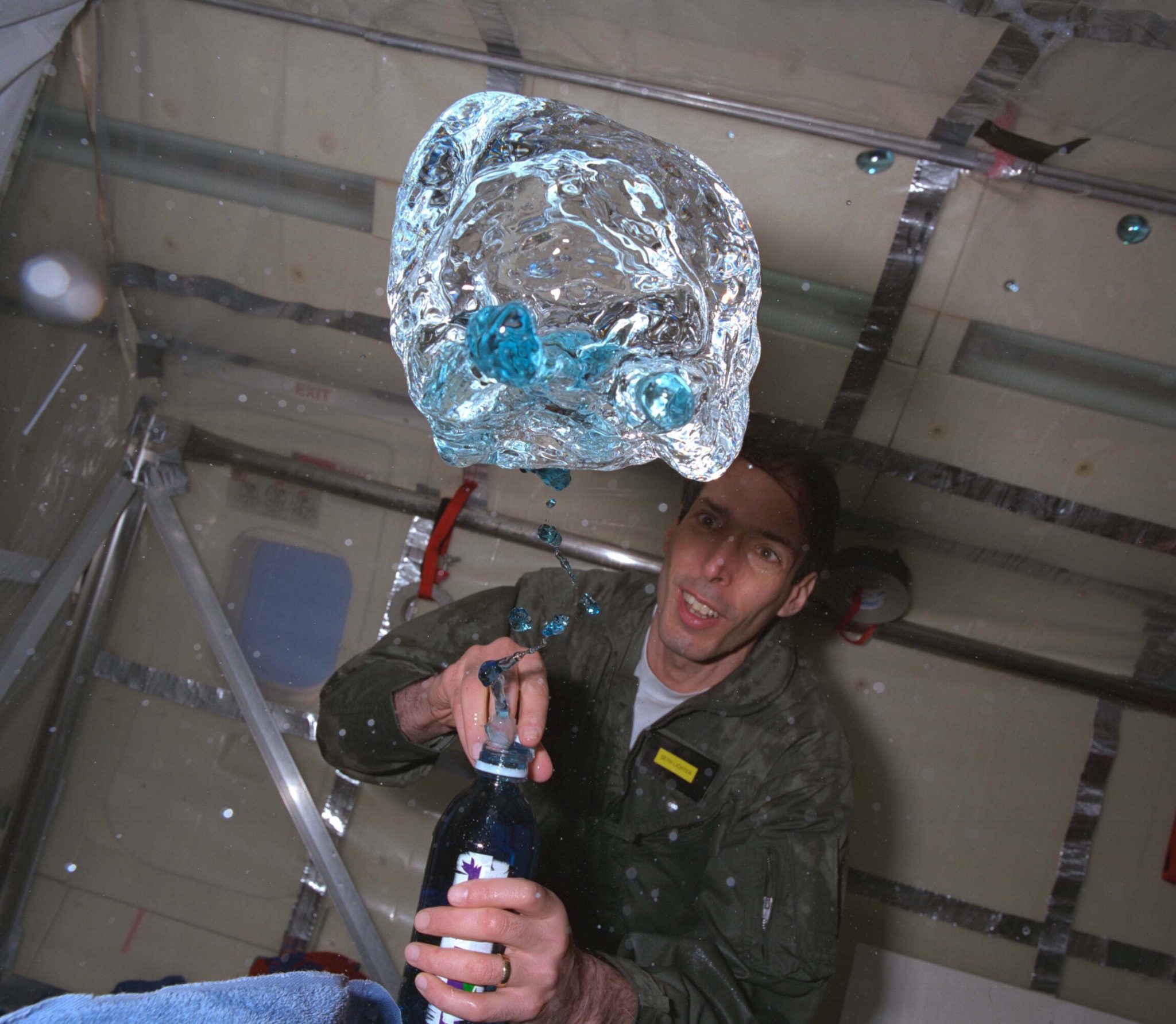 A person in a green flight suit watches as blue fluid floats out of a bottle during a microgravity flight.