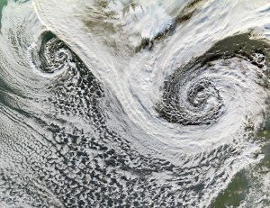 Two cyclones swirl south of Iceland in this satellite image. While the two storms' distinctive spiral patterns are clearly visible at left and right, white clouds cover most of the image. Some of Iceland can be seen at the top middle of the image, as well as part of Scotland at bottom right.
