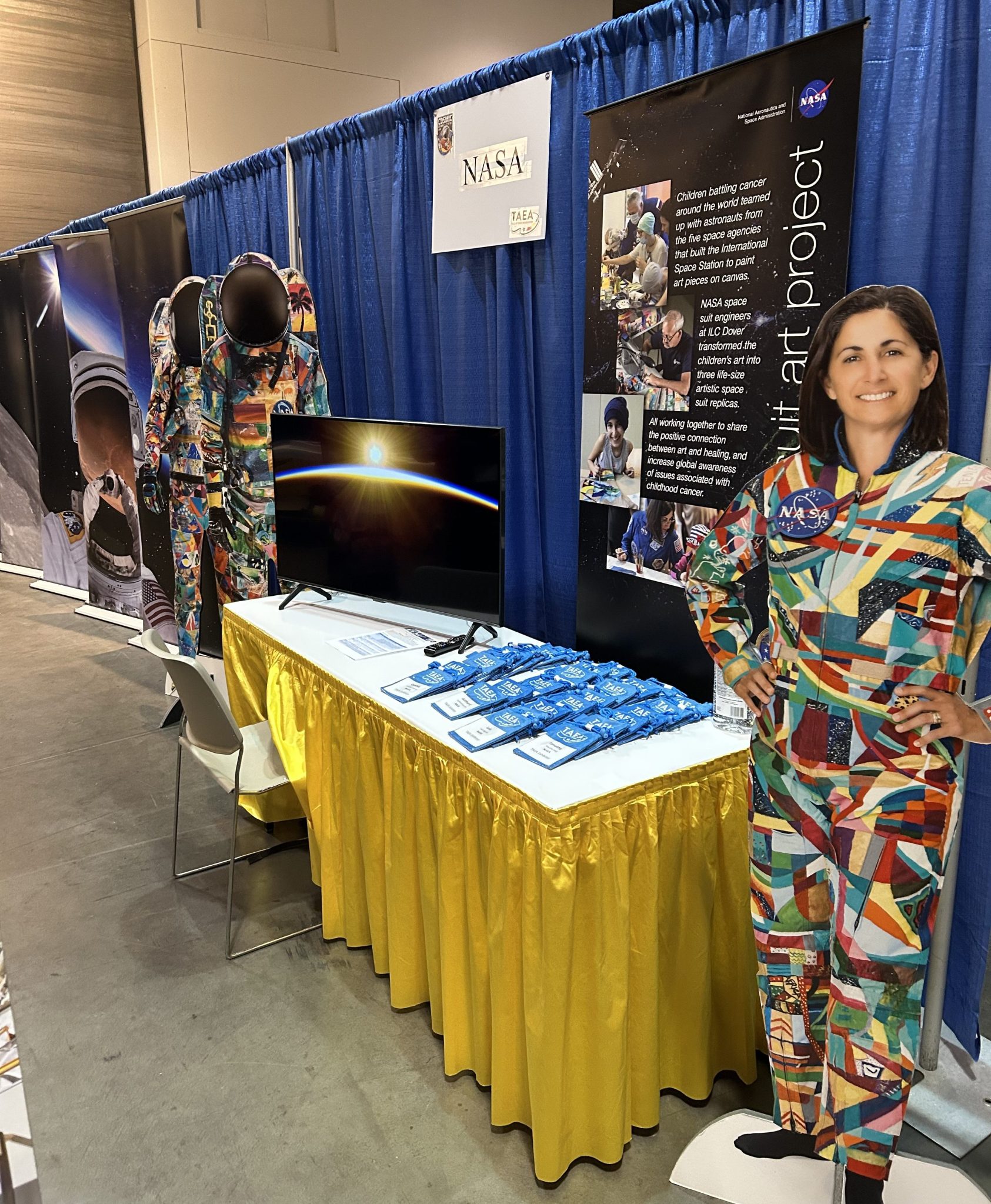 A NASA booth display features a table with a yellow skirt, showcasing informational materials. Behind the table, colorful artwork-themed astronaut suits and a life-size cutout of a smiling individual wearing a vibrant patchwork suit add visual interest. A TV screen displays an image of the sunrise from space.