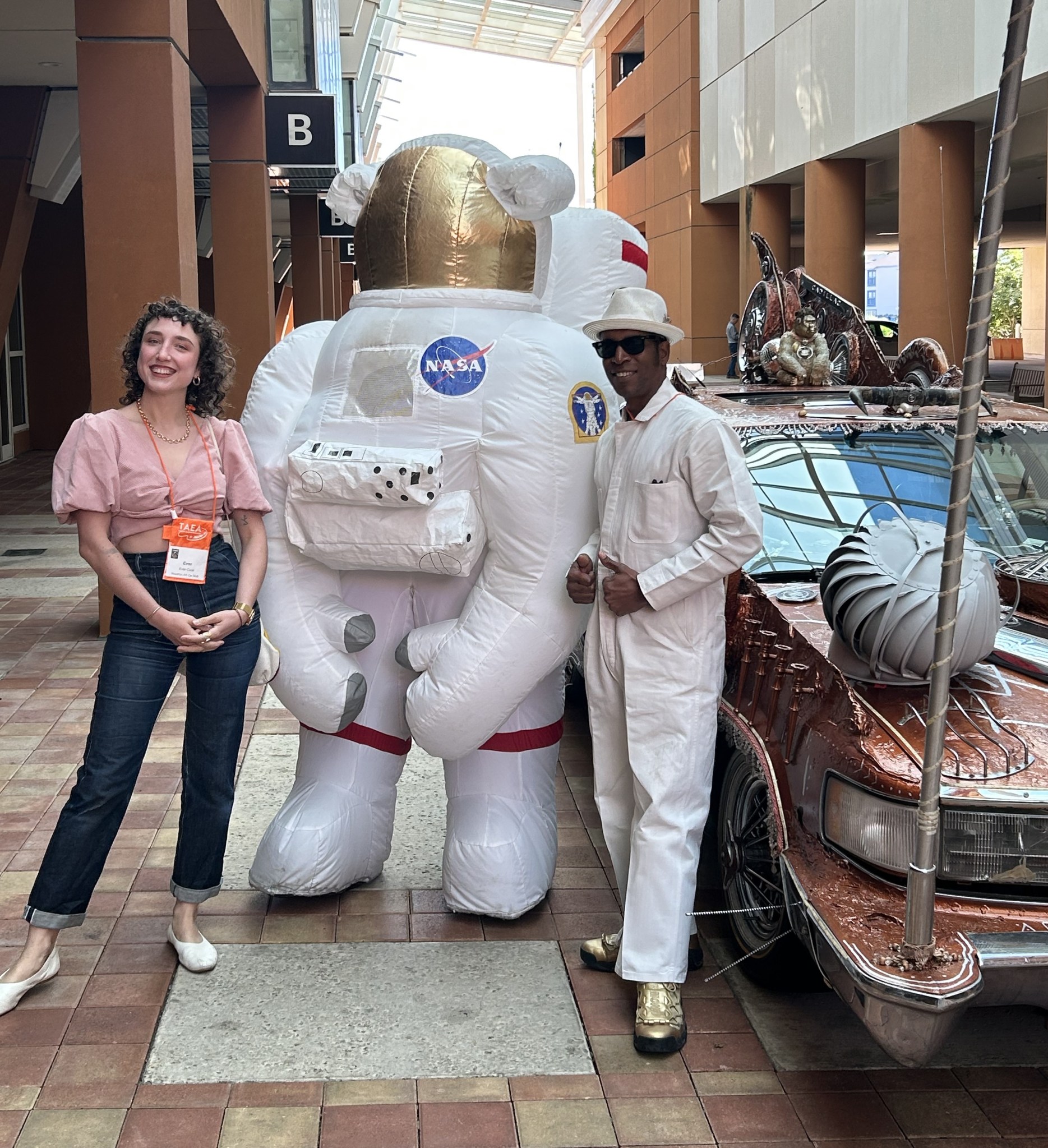 Two individuals stand on either side of an inflatable NASA astronaut suit. The setting is an indoor venue with high ceilings and modern architecture.