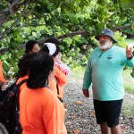 A Puerto Rican man with a bushy white beard, green bucket hat, teal longsleeve shirt, and black shorts gestures to a group of high school students wearing neon orange shirts.