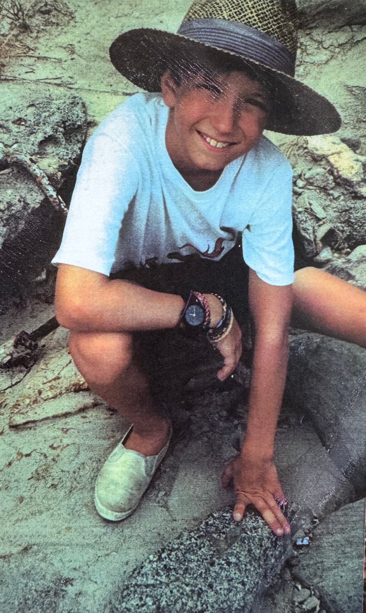 A young, smiling boy in a wide-brimmed hat crouches on some rocks.