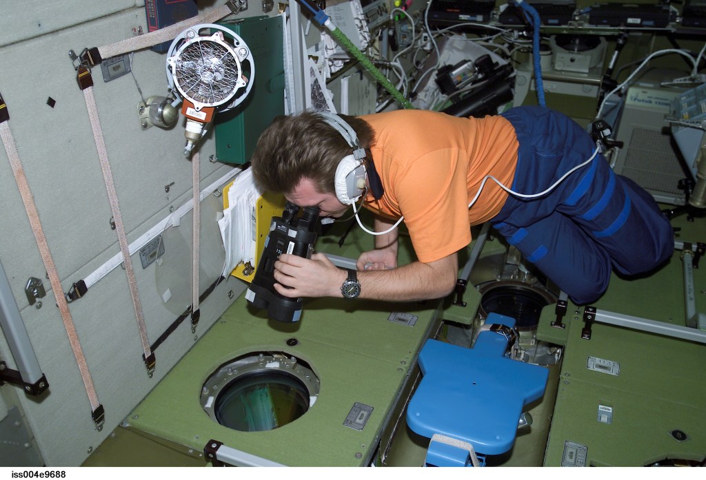 ISS004-E-9688 (24 March 2002) --- Cosmonaut Yury I. Onufrienko, Expedition Four mission commander, uses binoculars to look out a window in the Zvezda Service Module on the International Space Station (ISS). The crew anticipated the approach of a Progress supply vehicle about the time this image was taken.