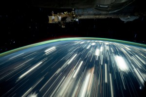 City lights on Earth appear as bright streaks in this long-exposure photo. Earth's atmosphere glows green on the horizon. Part of the International Space Station is visible at top right.