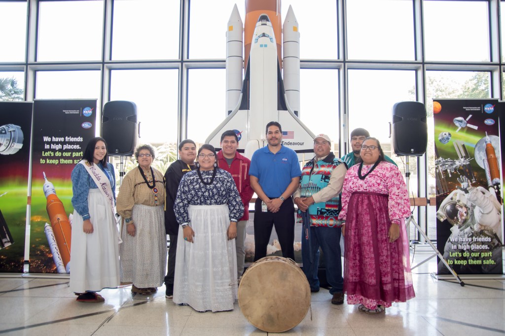 A group of Indigenous people and a NASA employee pose together in front of a model of the Space Shuttle Endeavour. The individuals wear traditional clothing, including sashes and beaded garments.