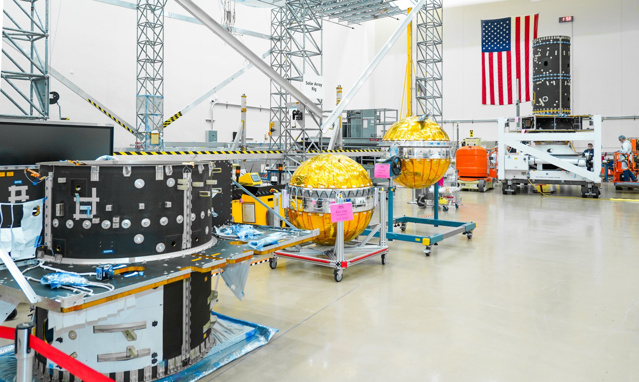 The Power and Propulsion Element (PPE) hardware for the Gateway space station, including its primary structure and gold-coated fuel tanks, is displayed in a cleanroom at Maxar Space Systems in Palo Alto, California. The components are organized for assembly, with an American flag visible in the background.
