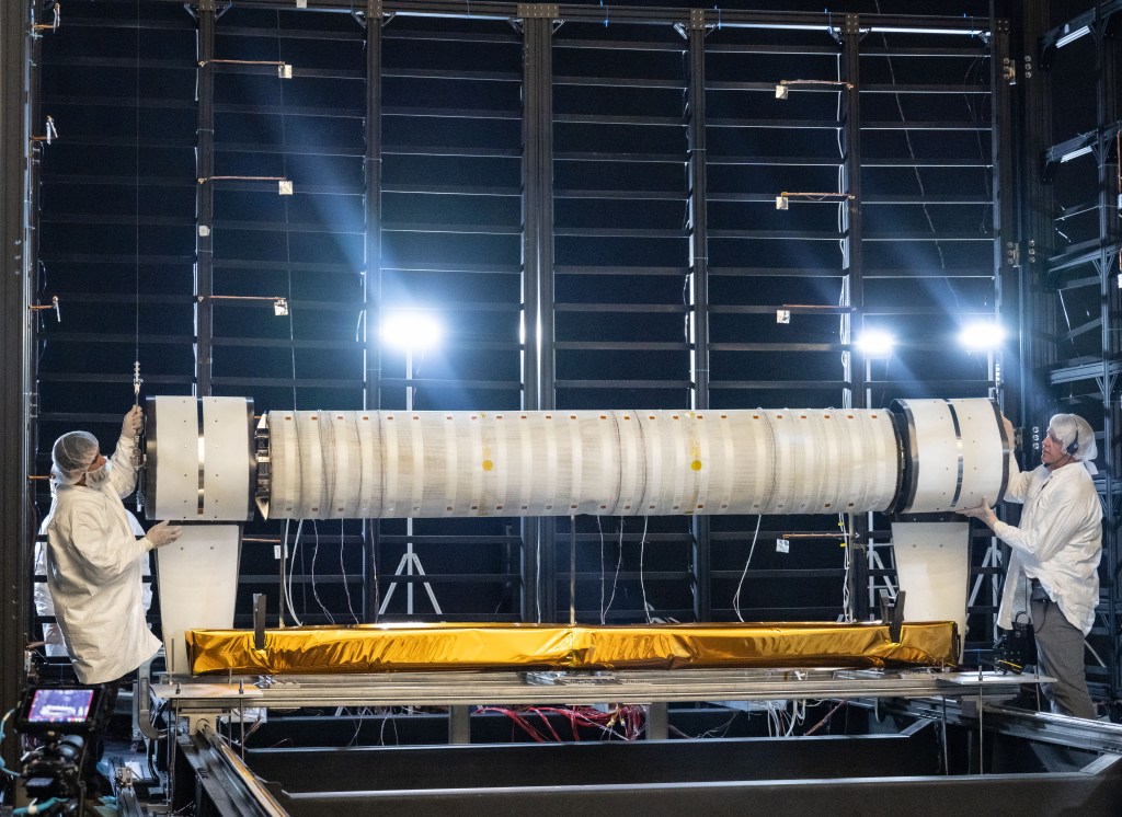 Two technicians in cleanroom attire work on a cylindrical component suspended horizontally inside a testing chamber.