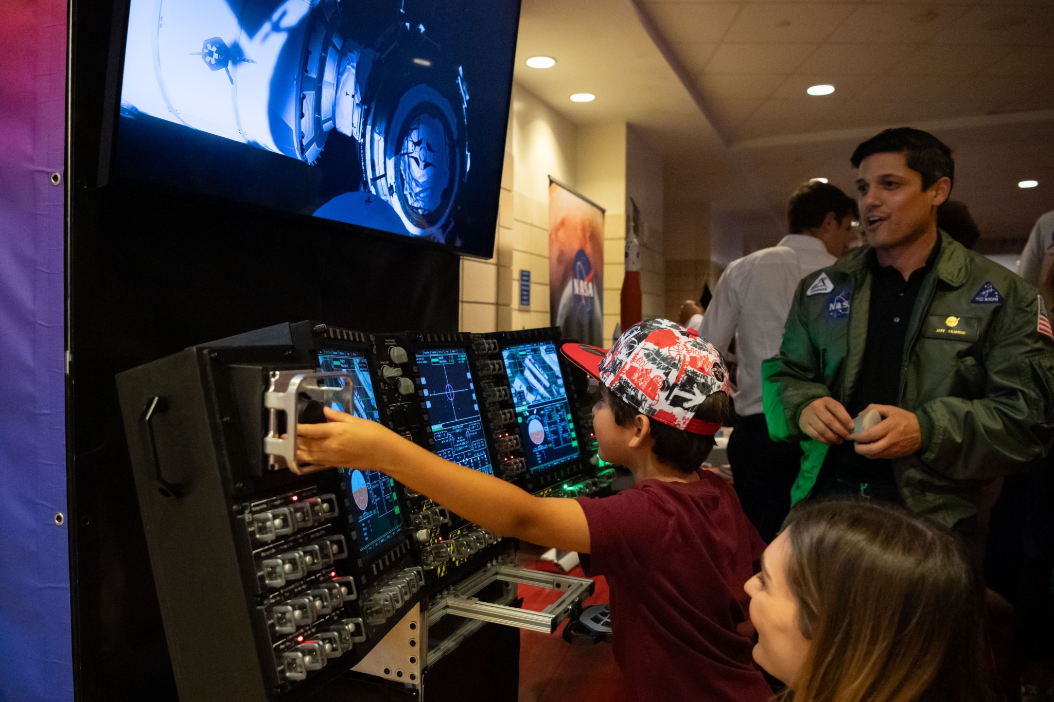 A young child wearing a colorful cap interacts with a NASA control panel simulator while an adult in a green NASA flight jacket observes nearby.