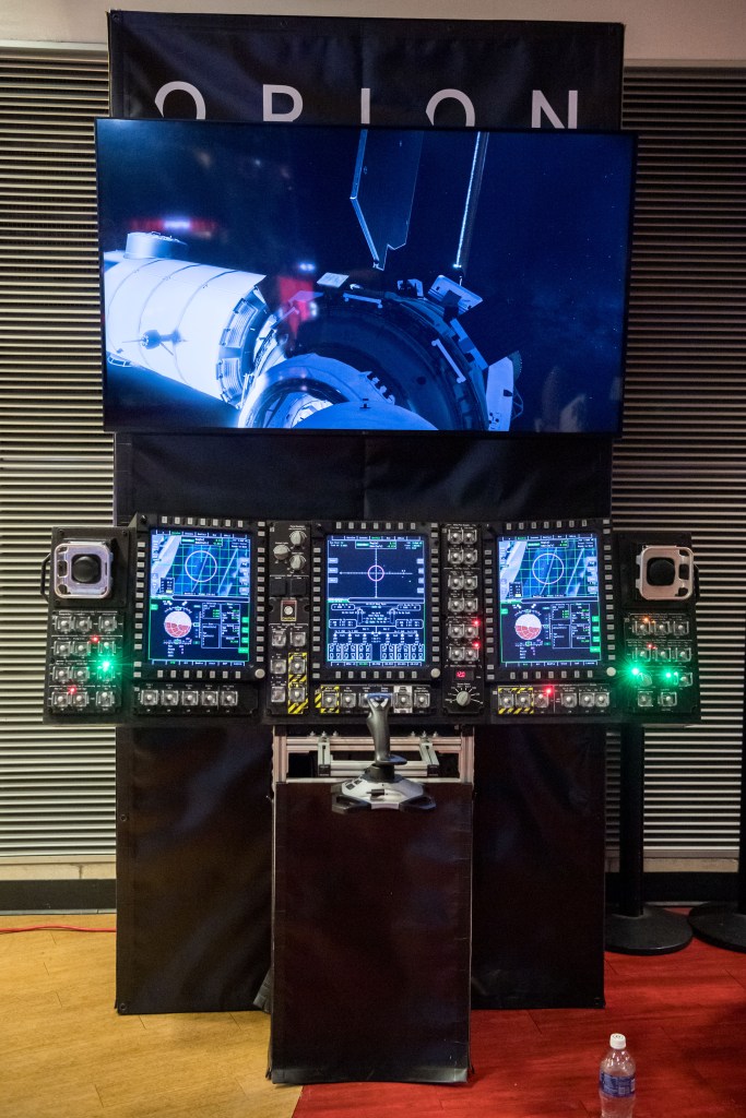 A detailed view of a NASA Orion spacecraft control panel simulator, featuring multiple screens displaying spacecraft navigation and docking data. 