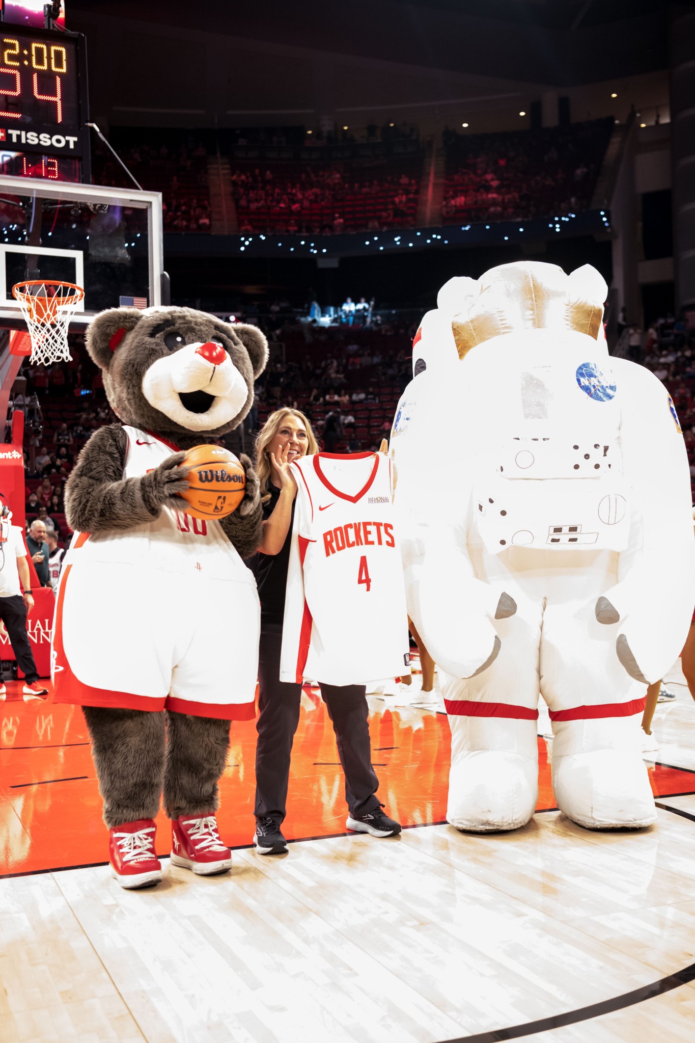 A woman poses on a basketball court holding a Houston Rockets jersey with the number 4. She is joined by the Houston Rockets mascot, a bear holding a basketball, and an inflatable NASA astronaut mascot. 