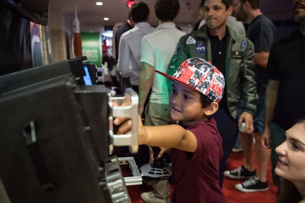 A young boy wearing a red and white patterned cap interacts with a NASA control panel simulator, smiling as he holds the joystick. A woman kneels beside him while a man in a green NASA flight jacket observes in the background. 