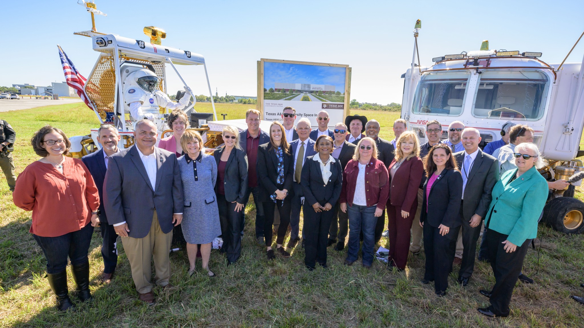 A group of attendees gathers for a group photo at the Texas A&M University Space Institute event, standing in front of a space exploration rover and a conceptual rendering of the institute building.