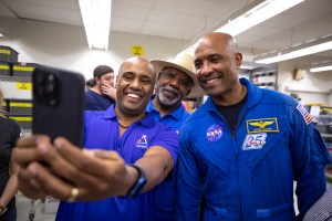 Three African American men - two Kennedy Space Center employees wearing blue polo shirts with an Artemis logo (left, middle back) and NASA astronaut Victor Glover (right) - pose for a photo taken with a cell phone.