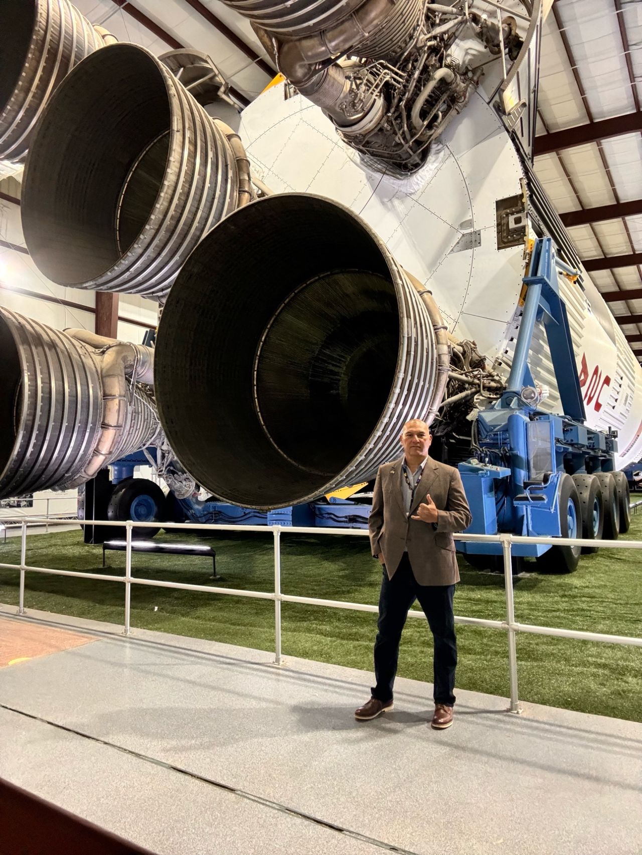 A man stands in front of large rocket engines inside a hangar, giving a thumbs-up.