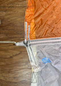 6)A sample of a Main parachute canopy at the skirt. This shows the area where the Kevlar radial webbing is joined to the Kevlar cord suspension line. Understanding the efficiency of this joint is critical to understanding the structural capability of the parachute. Also seen are metallic reefing rings that are used to allow the reefing lines to constrain the circumference of the skirt during inflations. These rings have since been replace with textile versions, akin to belt looks on a pair of inside-out jeans. Credit: Courtney Beasley/Jared Daum