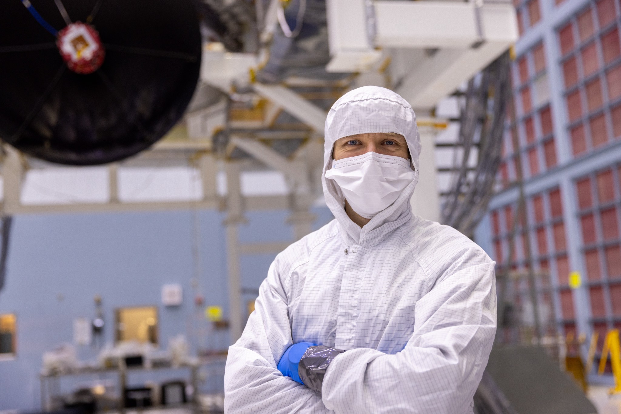 The upper half of a person in a white jumpsuit with hood and surgical mask stands in front of out-of-focus metal columns and a black disk.