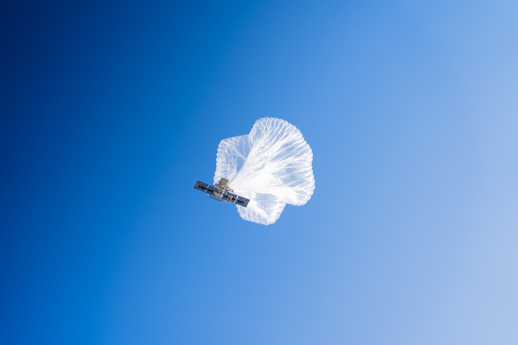 A high-altitude balloon is photographed from below as it ascends into a blue sky. The white balloon is partially inflated and the cell transmitter and other equipment is visible.