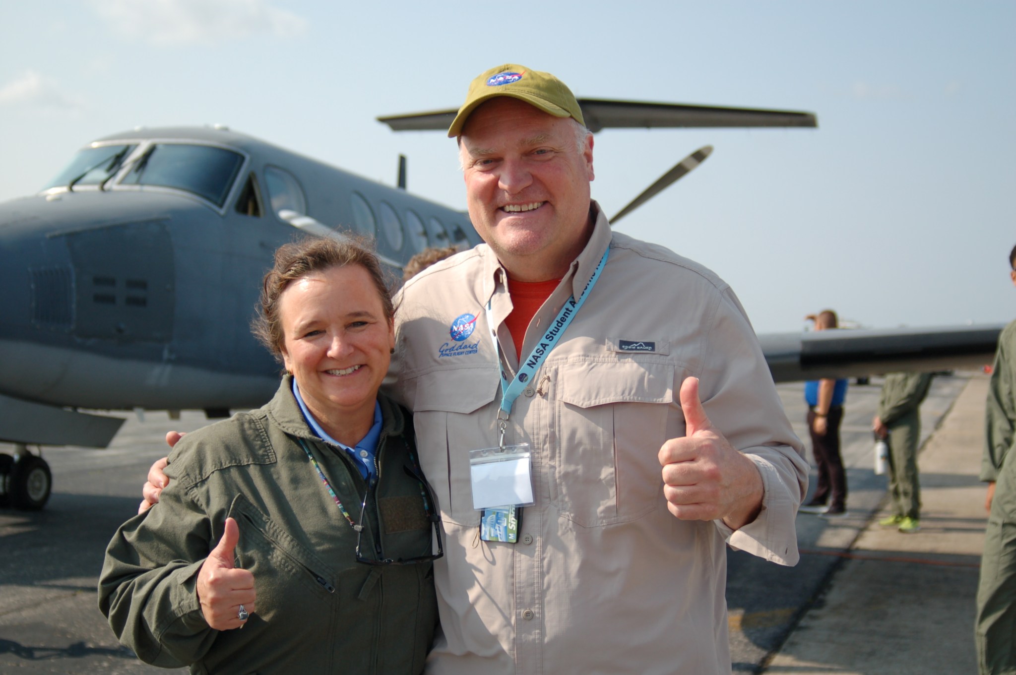 Woman wearing a green jacket and man wearing a tan button down shirt with the Goddard logo stand on a runway in front of a plane. 