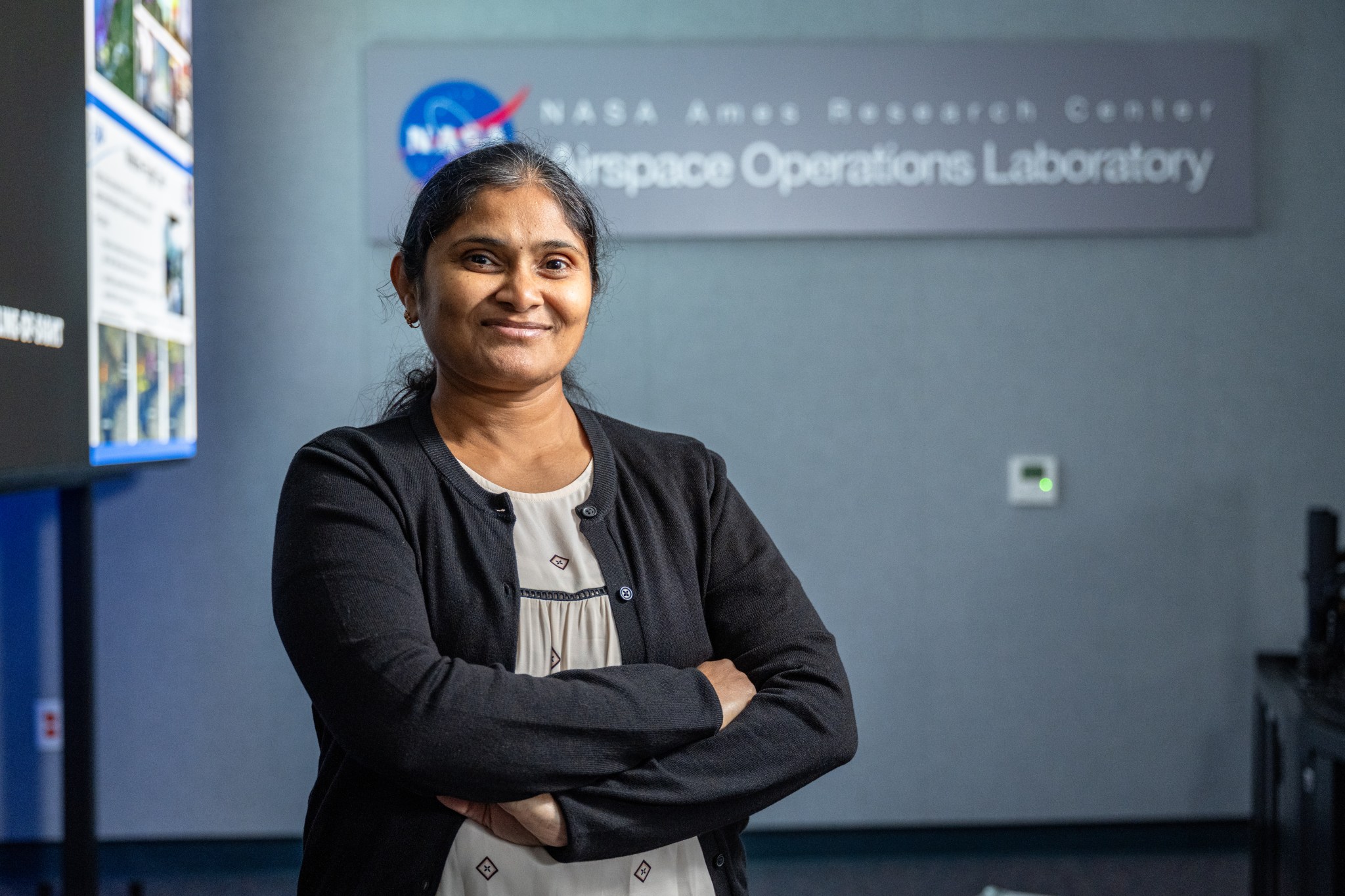 A woman wearing a white blouse with small black patterns on it and a black cardigan stands smiling with her arms crossed, in front of a sign reading "NASA Ames Research Center Airspace Operations Laboratory" and a large screen, which is blurred, but displaying text and images.