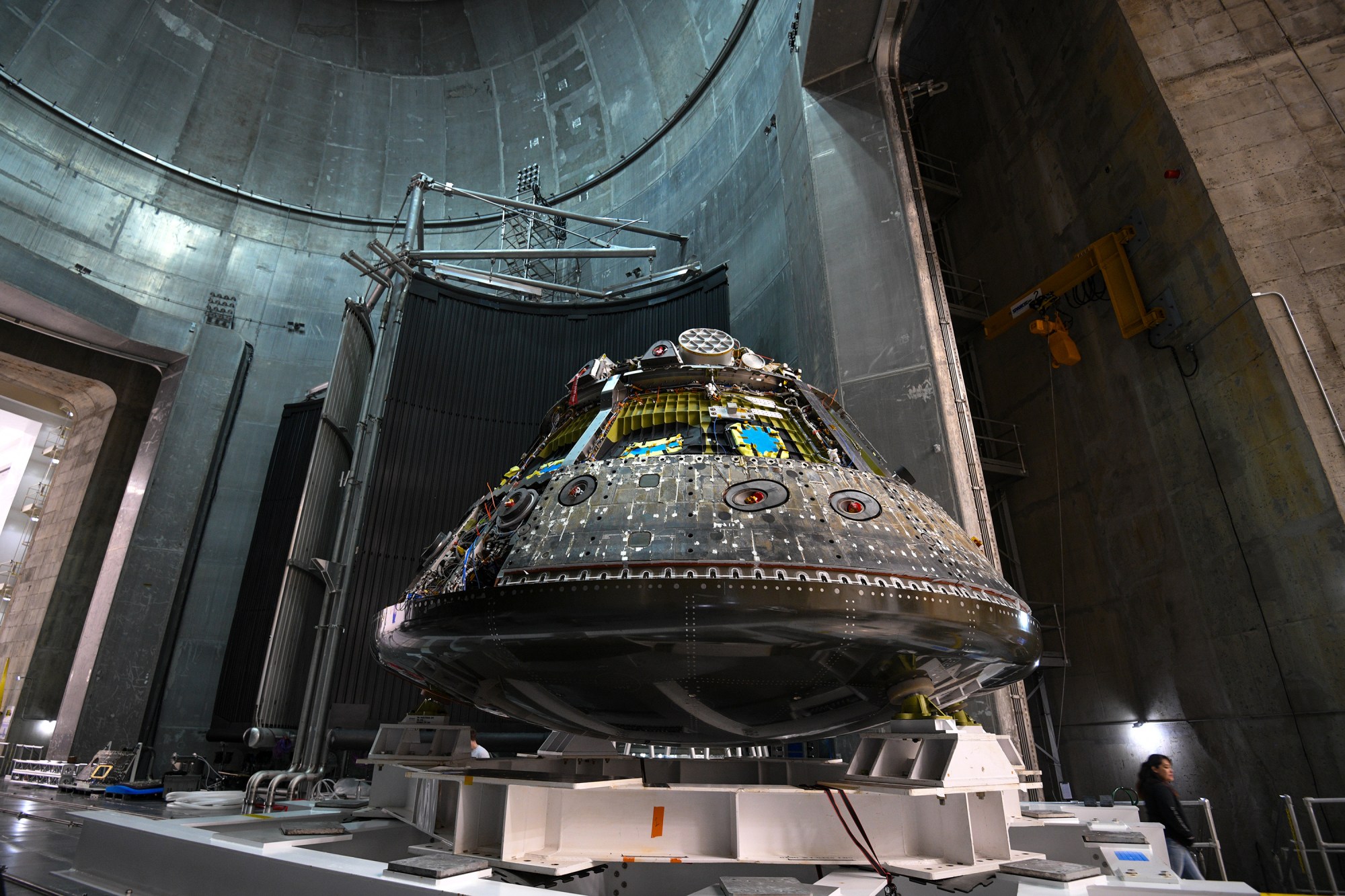 The Orion Environmental Test Article, blackened from its spaceflight, is photographed sitting on a white platform inside the shadowed aluminum vacuum chamber at NASA’s Neil Armstrong Test Facility. A woman with dark hair wearing a black sweatshirt appears in the lower right corner of the photo.