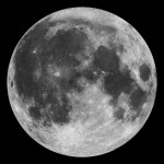 The full Moon stands our against the blackness of the night sky. The Moon appears as a white and gray orb dimpled with craters and valleys.