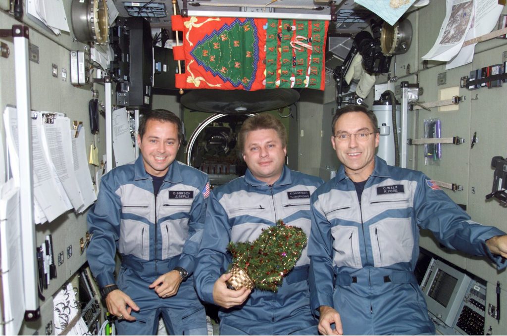 Three men in blue/gray suits smile as they pose for a Christmas pose onboard the Interrnational Space Station
