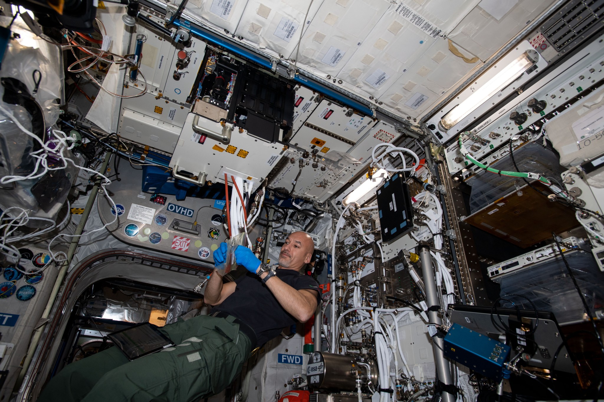 An astronaut aboard the International Space Station works with cables and tools while floating in a module filled with equipment, wires, and storage containers. The astronaut is wearing gloves, a black T-shirt, and green pants.